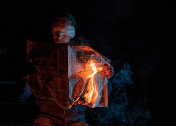 Blindfolded man reading a burning newspaper